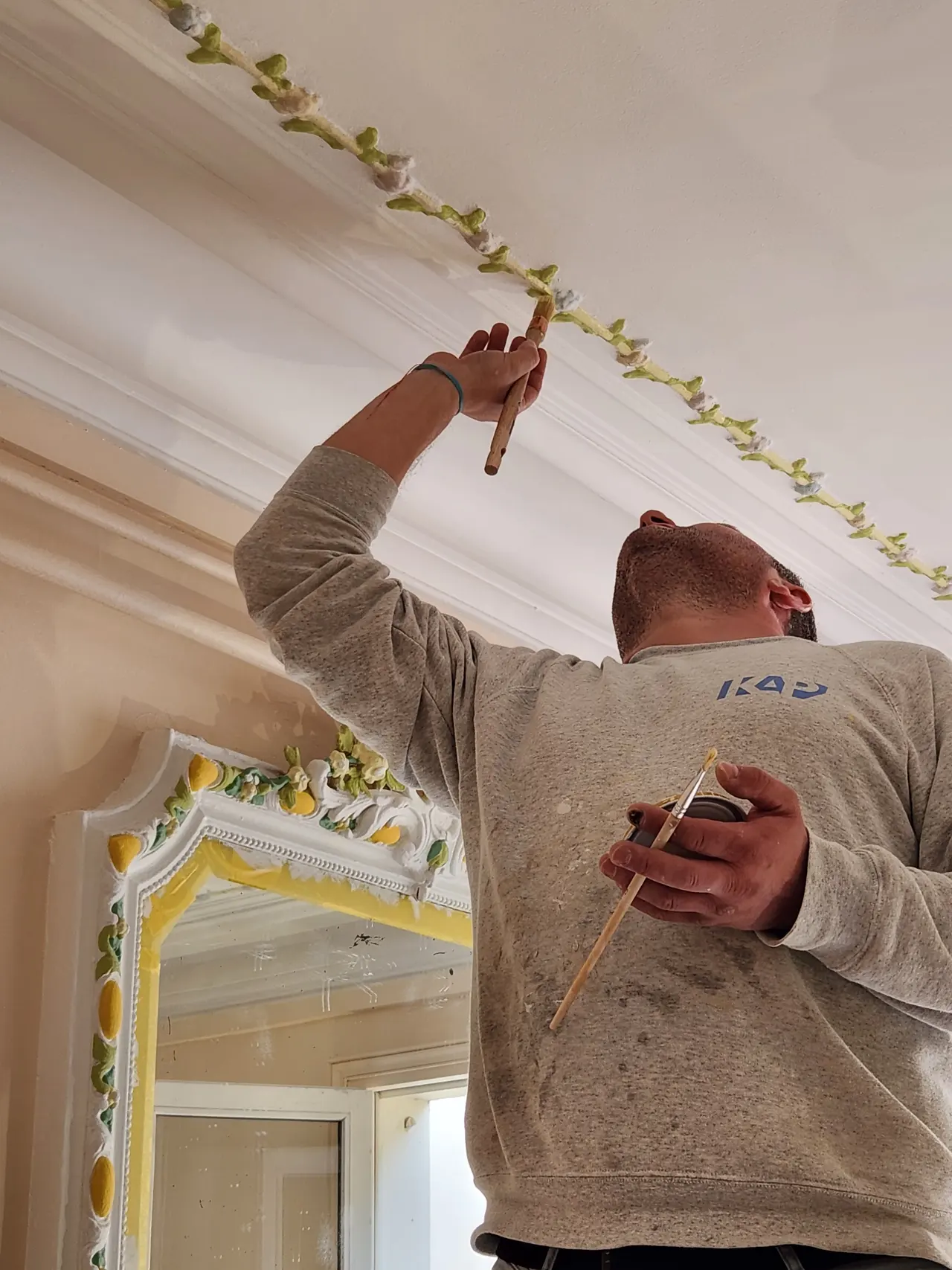 Un peintre retouche une frise peinte au plafond.